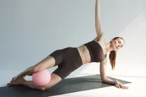 Esercizio di Pilates post parto: Carlotta Gagna nel plank laterale su un tappetino con una palla da Pilates sotto le gambe e un braccio teso verso l'alto, per rinforzare core e obliqui, nel programma Pilates Diastasi e Post Parto