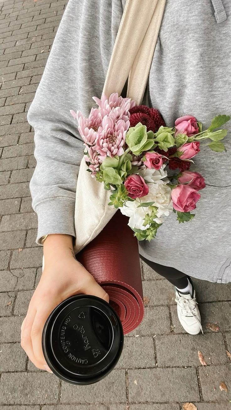 Donna che cammina con una borsa sulla spalla, un mazzo di fiori, un tappetino da Pilates e un caffè