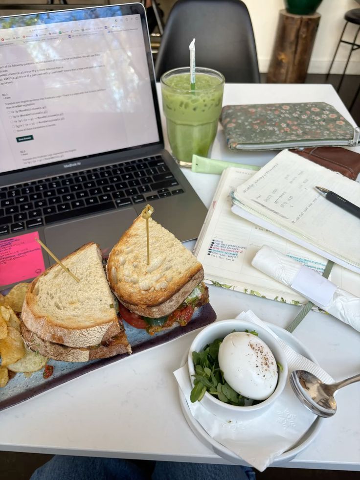 Scrivania di lavoro con computer aperto, appunti sparsi, un panino e un frullato
