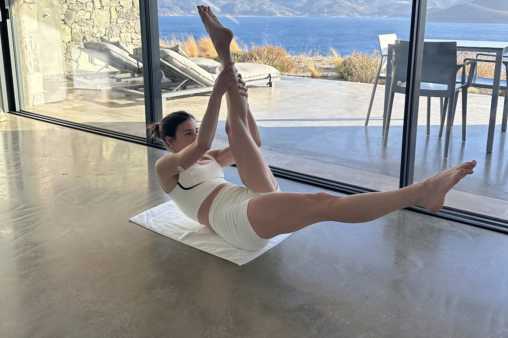 Carlotta Gagna esegue l'esercizio scissors del Pilates Linfodrenante® su un tappetino davanti alla vetrata con vista mare